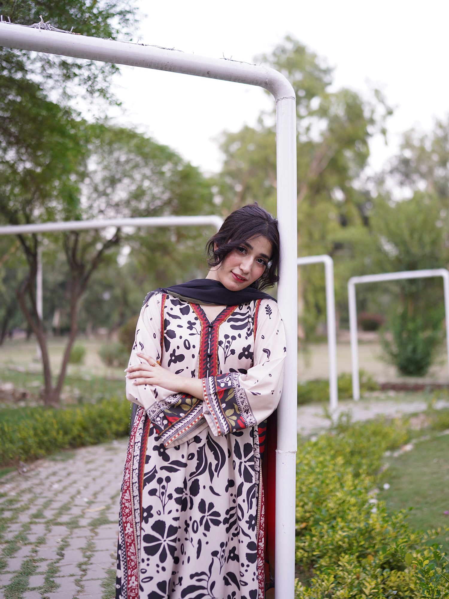 A woman wearing a black and white floral printed kurta with red and gold border details and a black dupatta, posing outdoors next to a white metal post in a park setting.