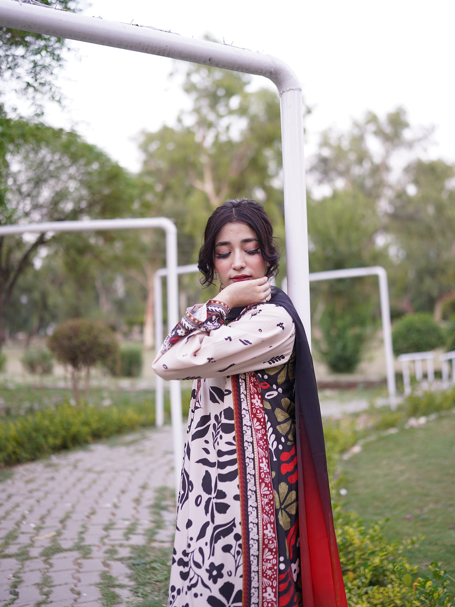 A woman wearing a traditional long-sleeve embroidered dress with a black, red, and beige floral pattern and a matching dupatta, standing in a park next to a white metal frame.