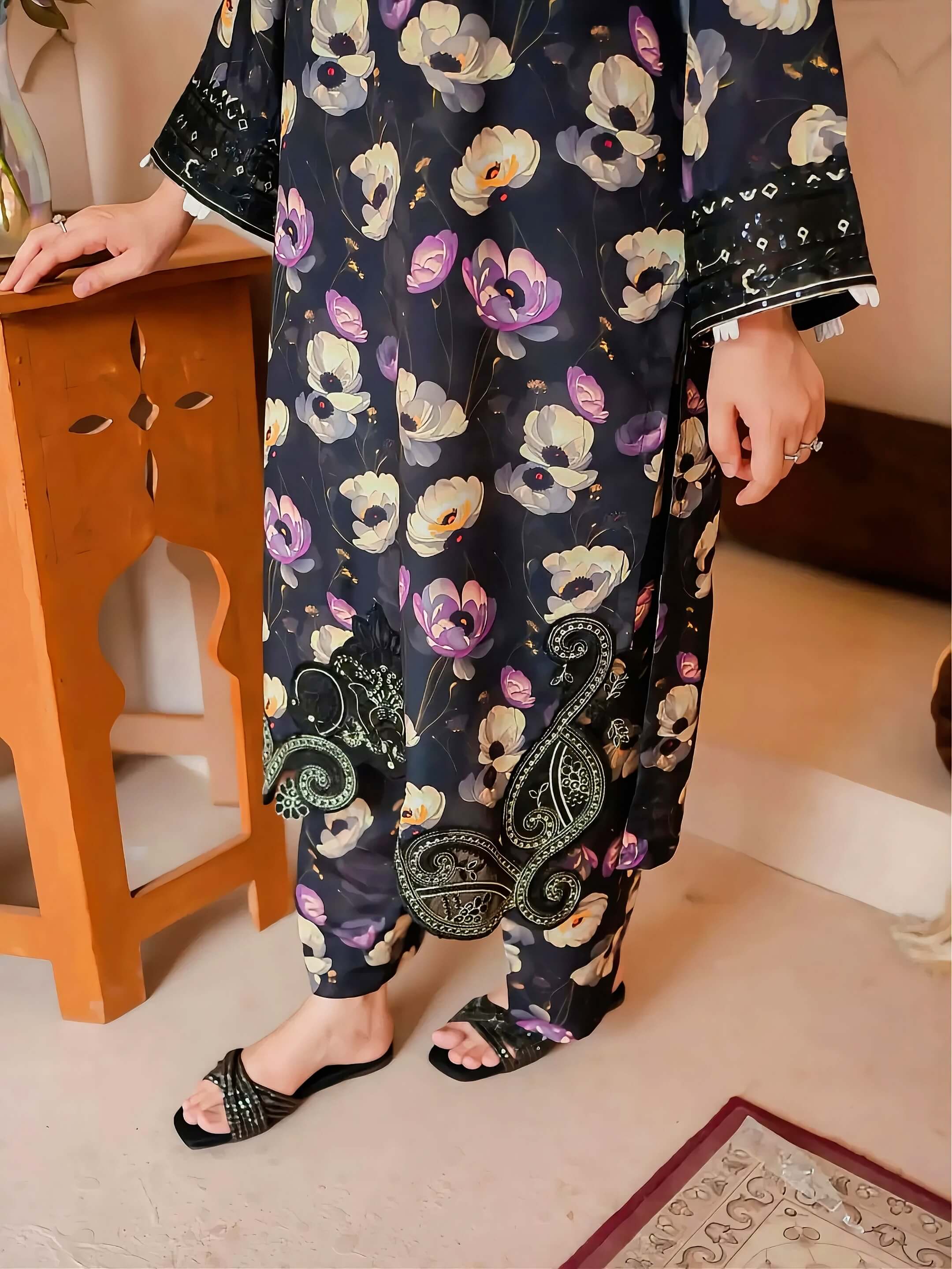 A person wearing a loose floral printed tunic and wide-leg pants set in dark navy, with large white, purple, and peach flowers and paisley embroidery near the hem, standing indoors next to a wooden table.