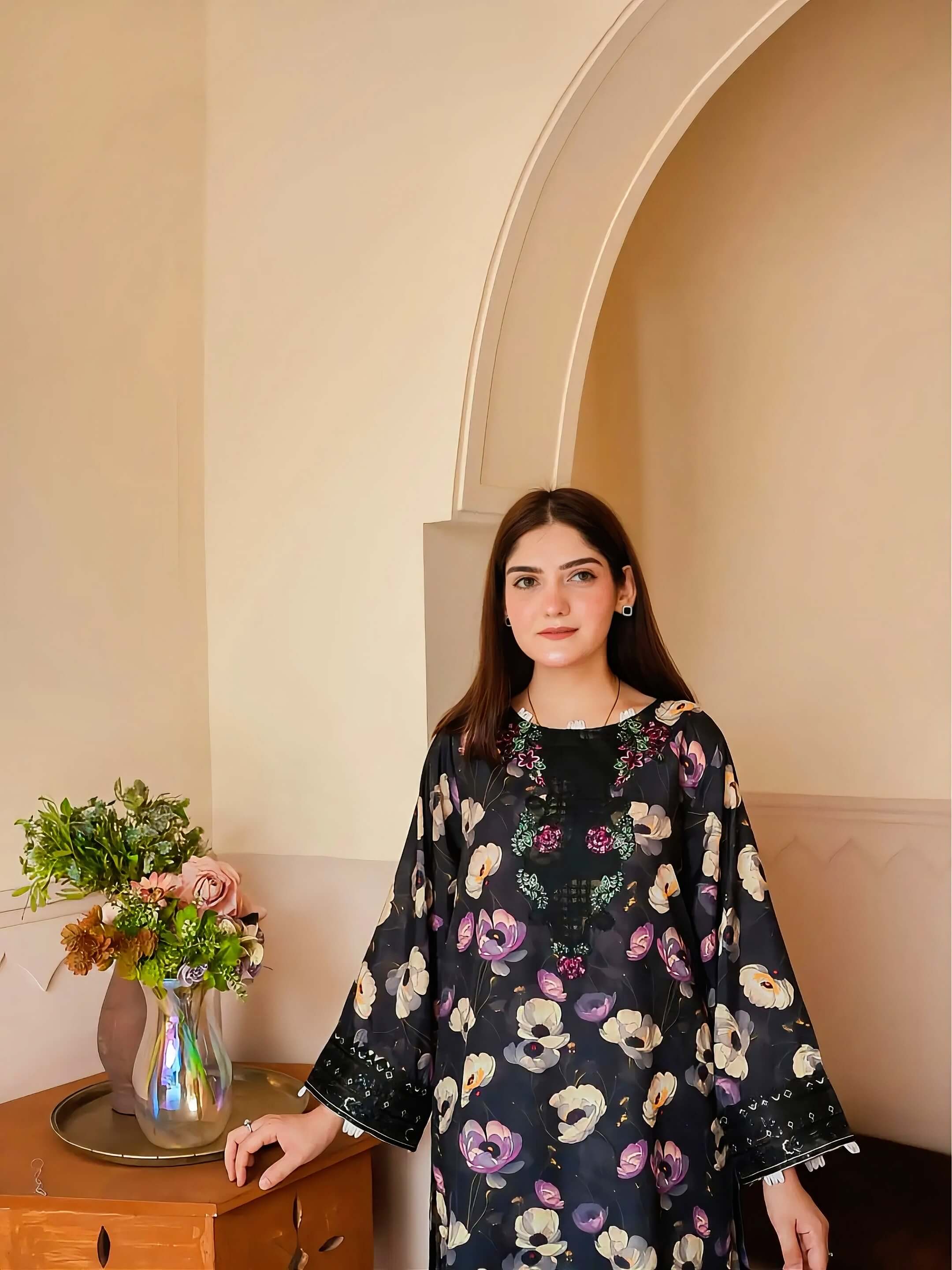 A woman wearing a black long-sleeve tunic with a colorful floral print, standing in an indoor setting with beige walls and a wooden side table holding a vase of flowers.