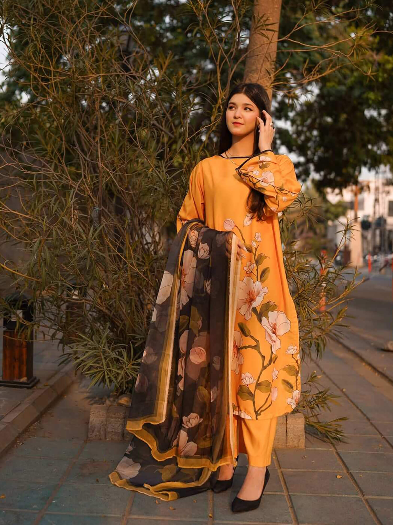 A woman stands outdoors on a paved walkway beside leafy trees, wearing a mustard yellow floral kurta with matching pants and a sheer black dupatta featuring large pink and beige floral prints with gold trim.