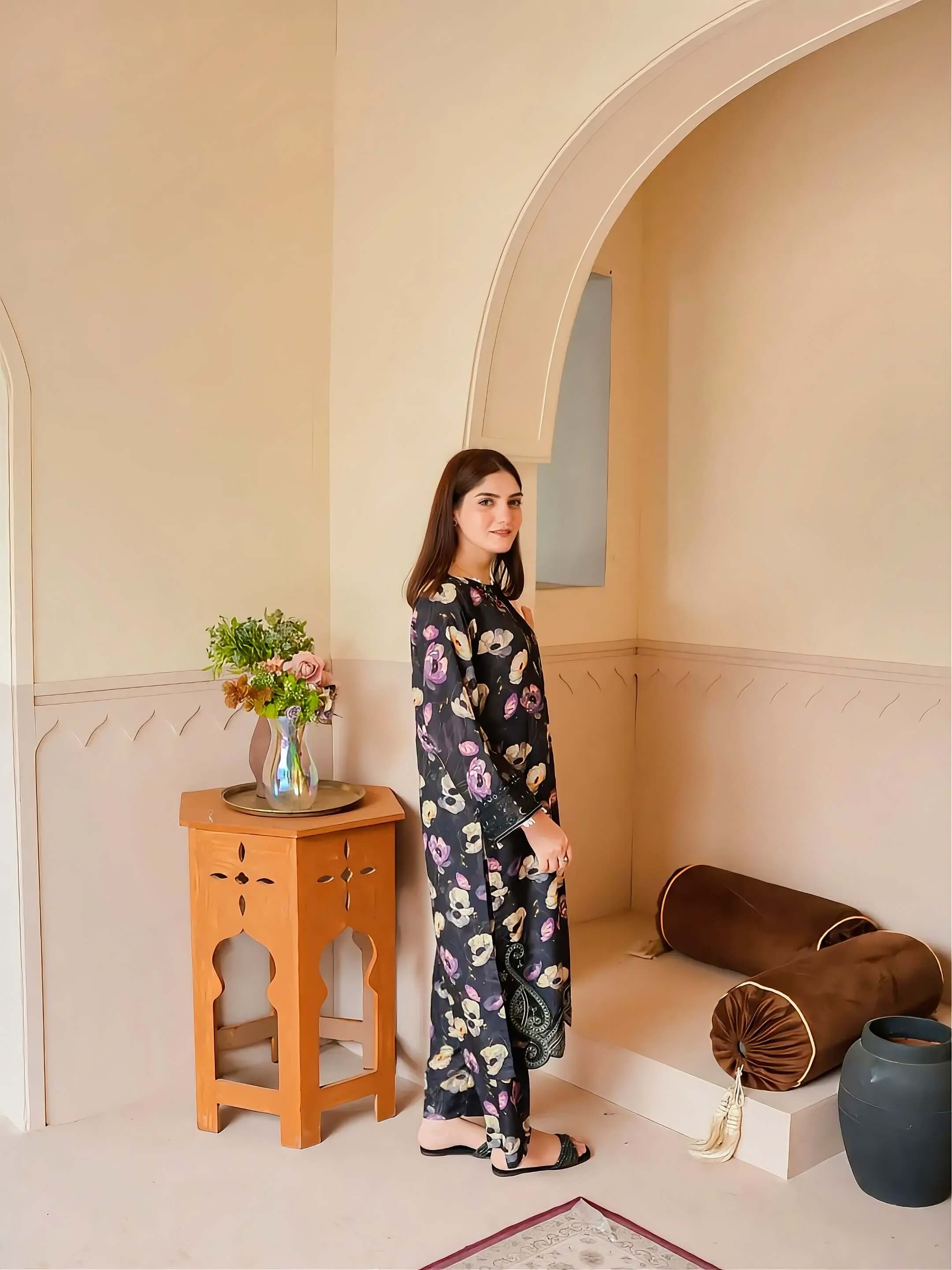 A woman stands in a softly styled interior wearing a long-sleeve, wide-leg kurta set with a dark base and a colorful floral print. The room features warm beige walls, arches, a small wooden side table with flowers, and decorative cushions.