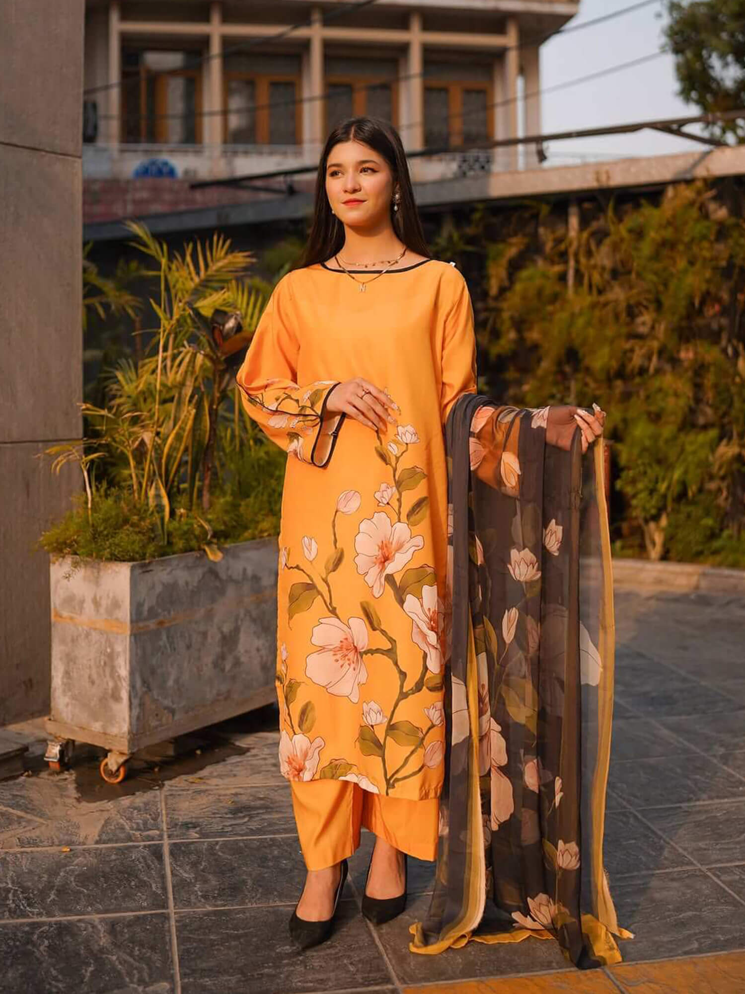 A woman models a bright orange-yellow three-piece salwar kameez suit with a floral print, holding a sheer dupatta with a gold border. The background shows a modern building and potted plants.