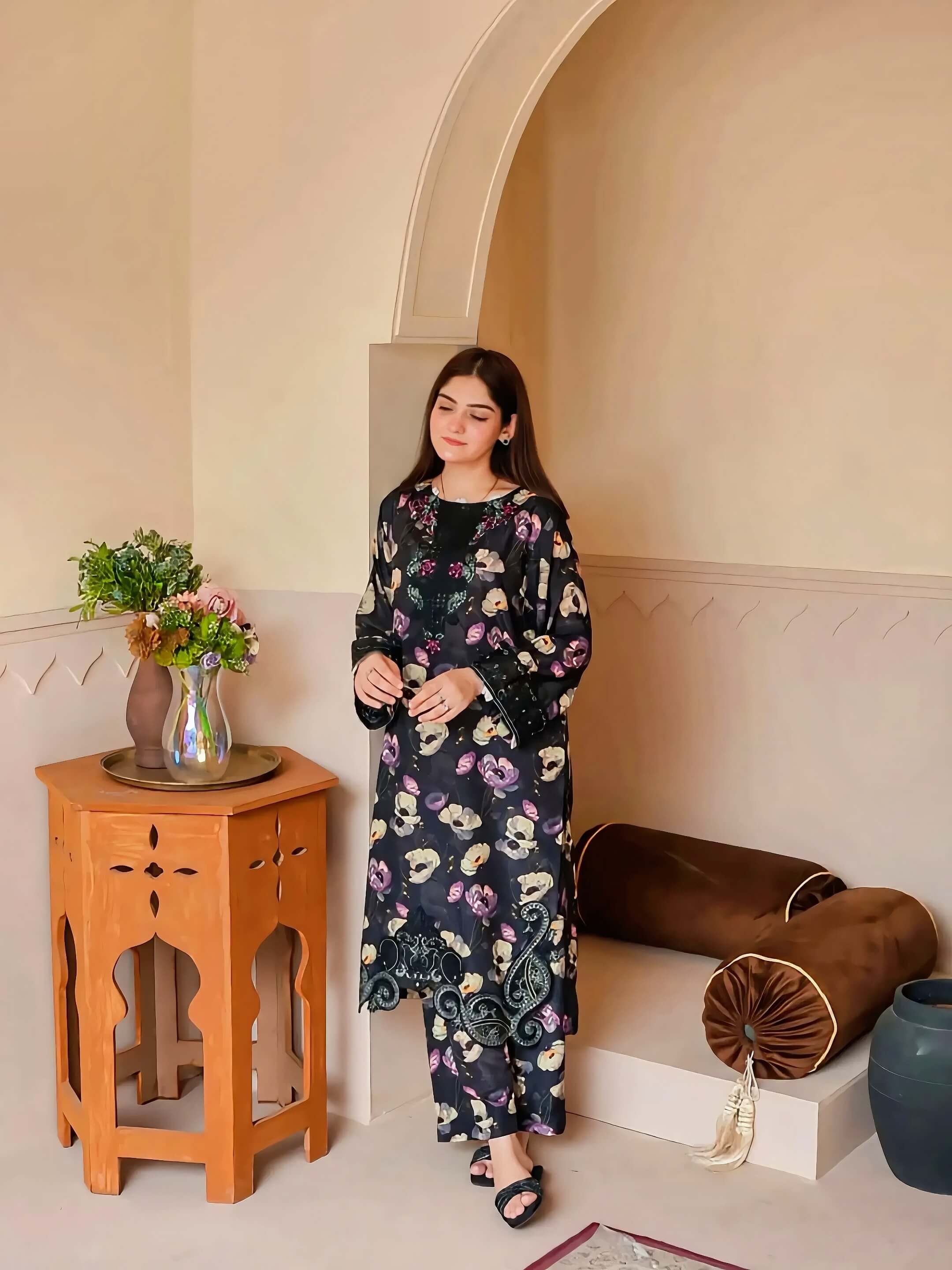 A woman models a black floral print kurta and matching pants set in a warm, beige-toned room with an archway, wooden side table, and cushions.