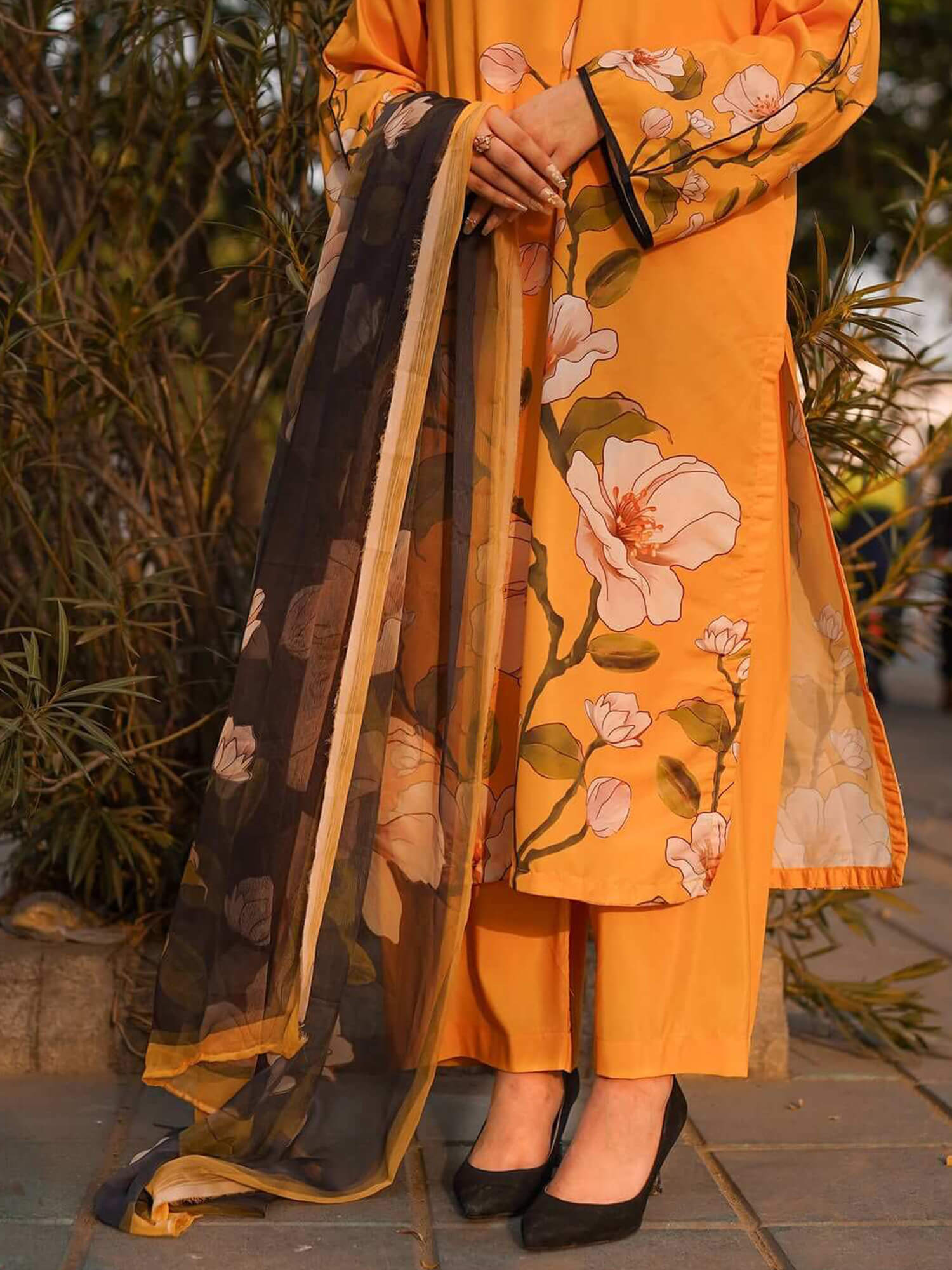 A woman wearing a mustard yellow floral printed kurta with matching pants and a sheer black dupatta featuring a gold border and floral print, standing outdoors on a paved surface with plants in the background.