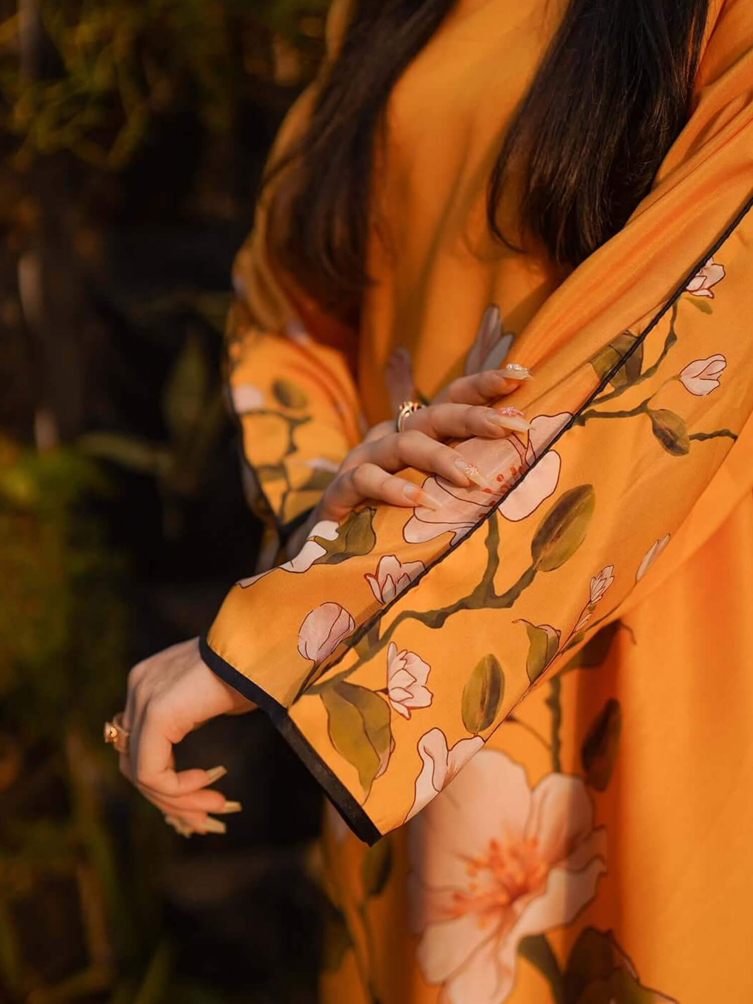 A close-up of a person wearing a flowing mustard yellow garment with a floral print in pink, white, and olive green, featuring black trim on the sleeve cuffs, set against a dark, blurred background.