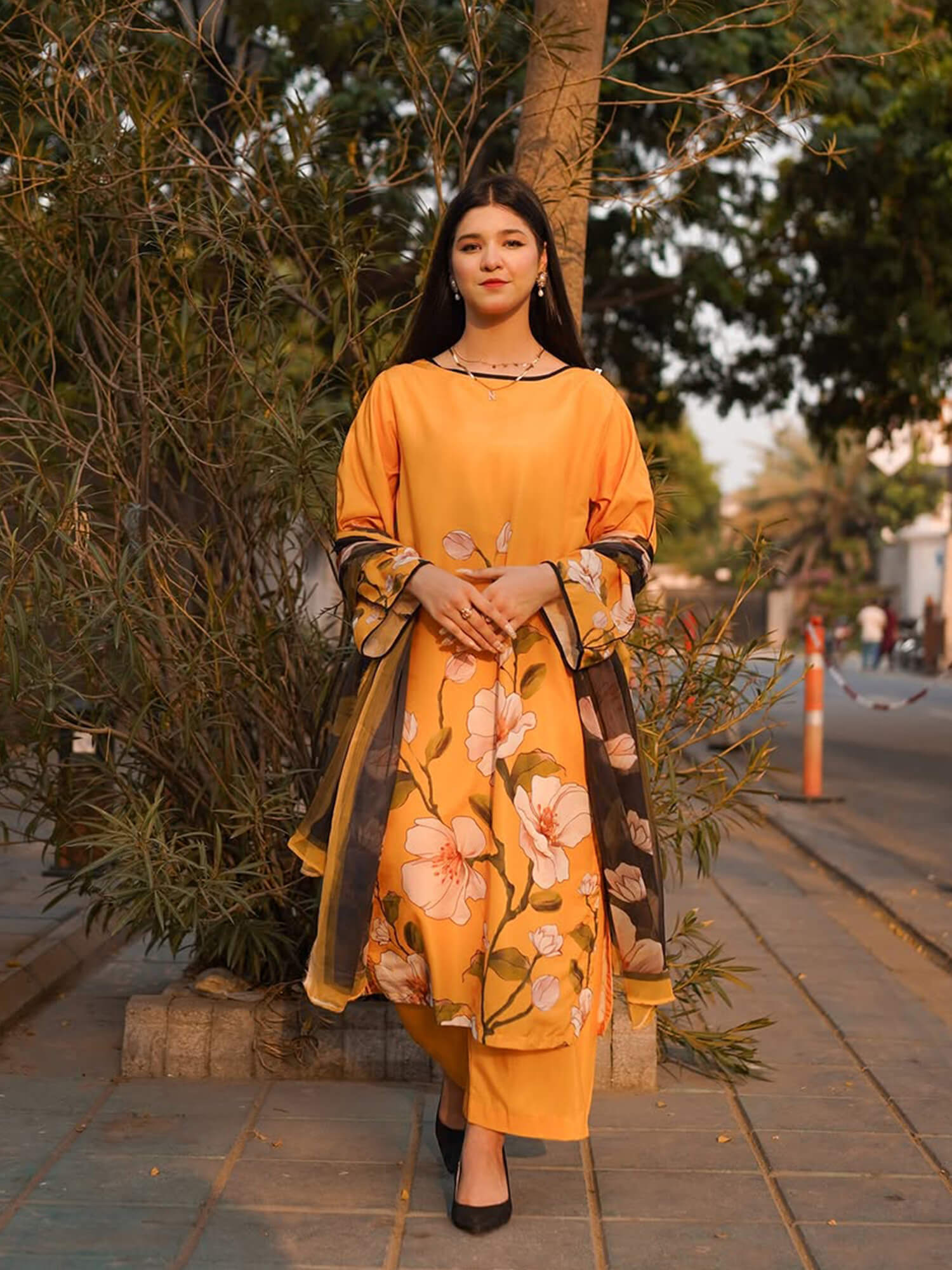A woman stands outdoors on a paved walkway wearing a mustard yellow kurta set with pink floral prints and a sheer black dupatta with gold border detailing.