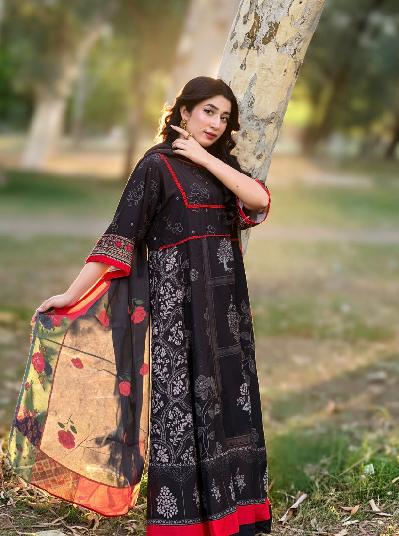 A woman wearing a black printed salwar kameez suit with red and beige border details, holding a matching dupatta with floral patterns, posing outdoors next to a tree trunk.