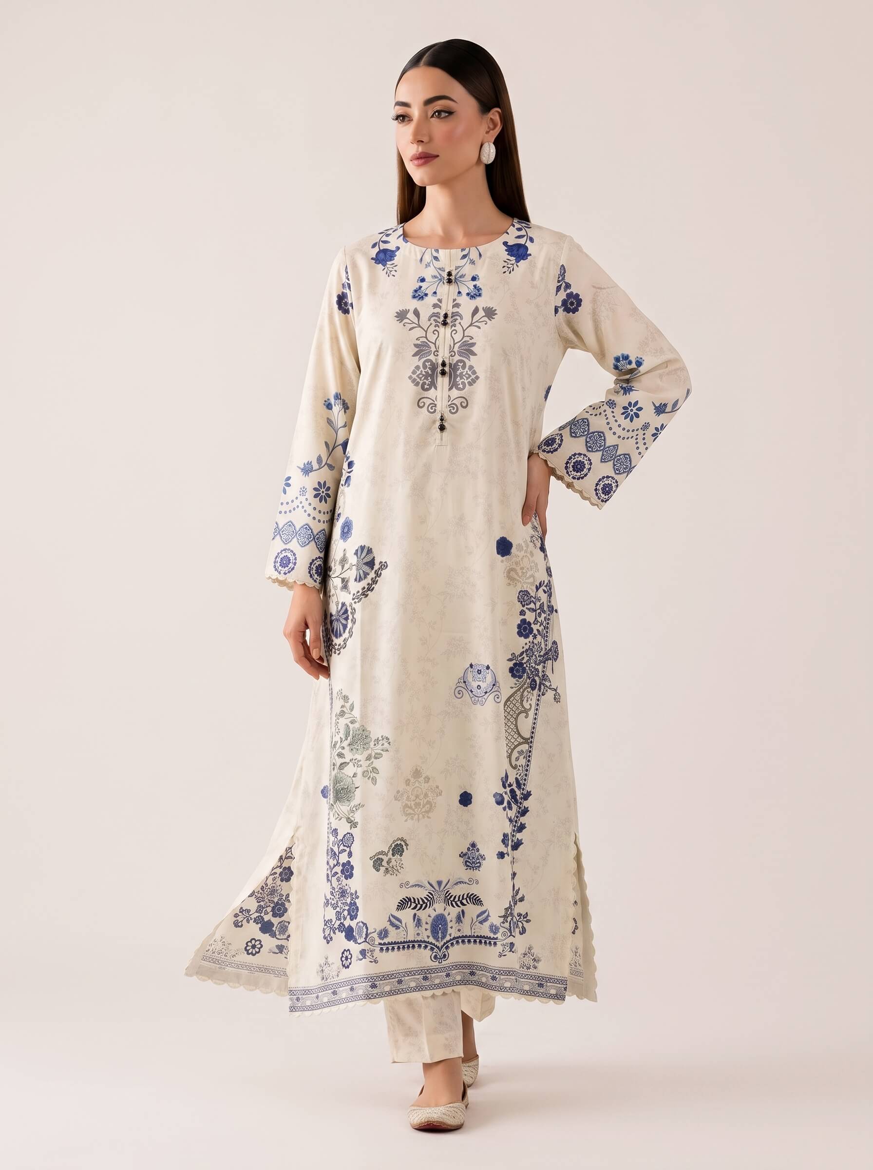 A woman modeling a long cream-colored embroidered kurta with blue floral and vine patterns, paired with matching cream pants, against a solid light beige studio background.