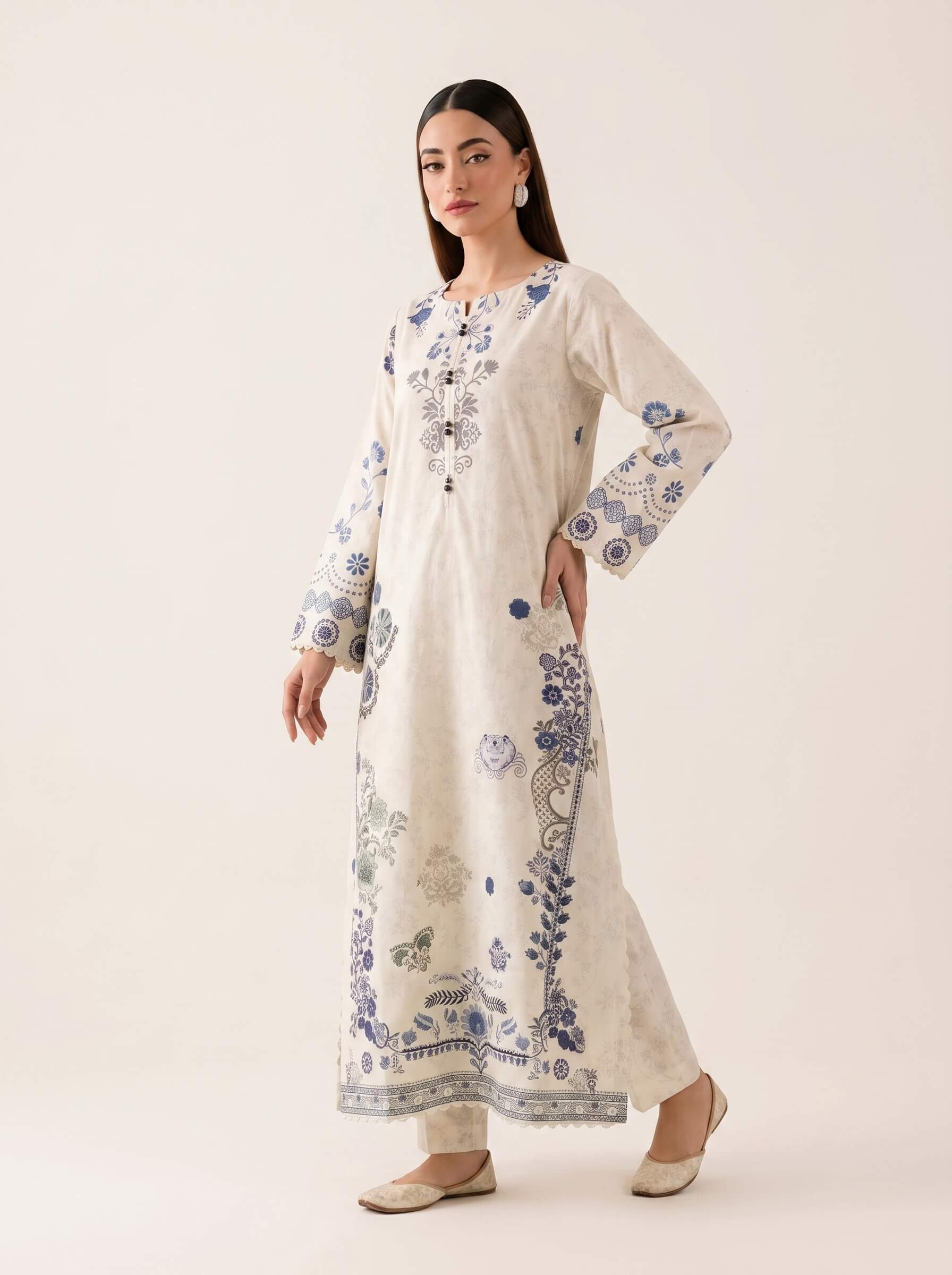 A woman modeling a long cream-colored kurta with blue floral and geometric embroidery, paired with matching cream trousers and beige flat shoes, against a solid light beige studio background.