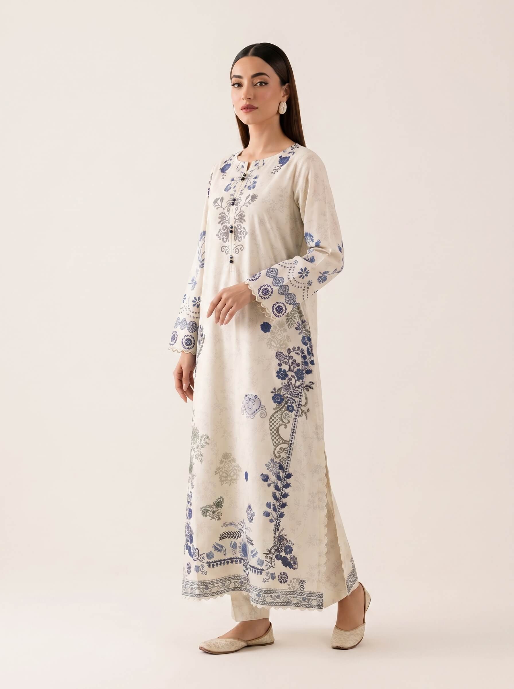 A woman modeling a long cream-colored kurta with blue floral and botanical prints, paired with matching cream trousers and beige flat shoes, set against a plain light beige studio background.