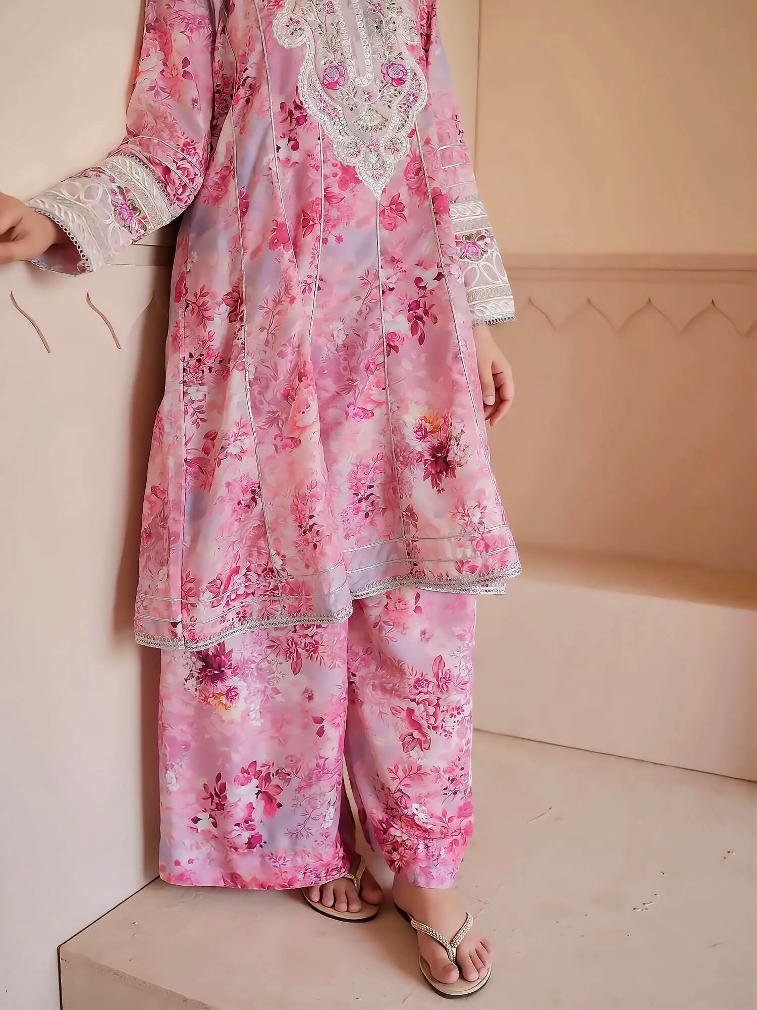 A person wearing a pink floral printed traditional outfit with matching wide-leg pants and white lace trim, standing indoors against a beige wall.
