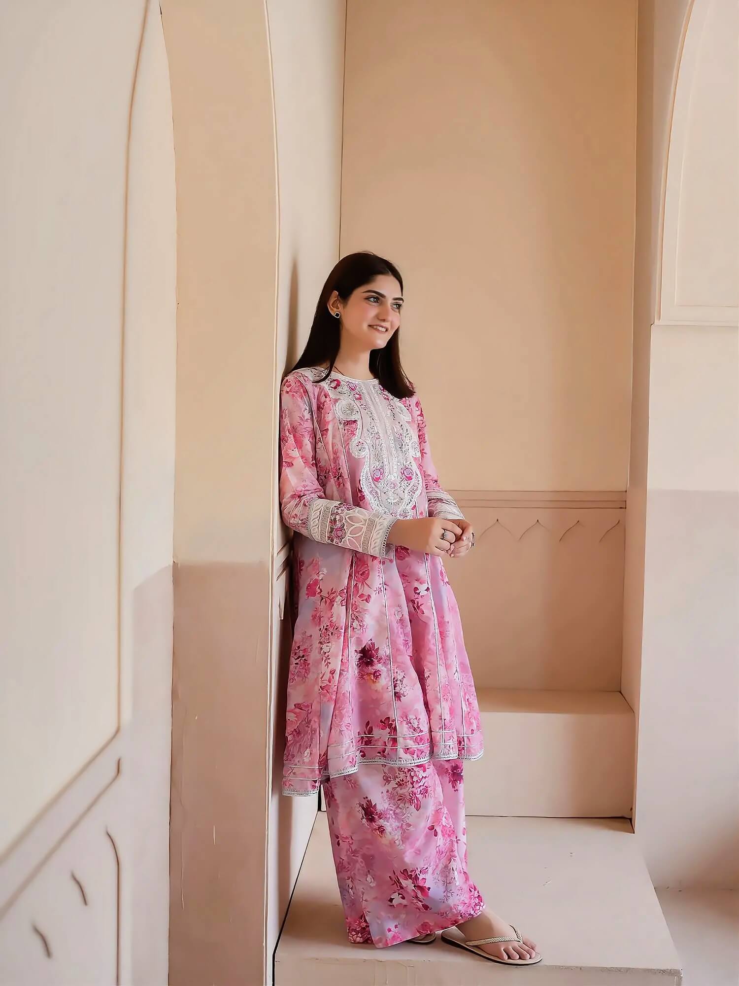 A woman wearing a pink floral printed kurta and matching pants with white lace trim, standing in a beige architectural setting.