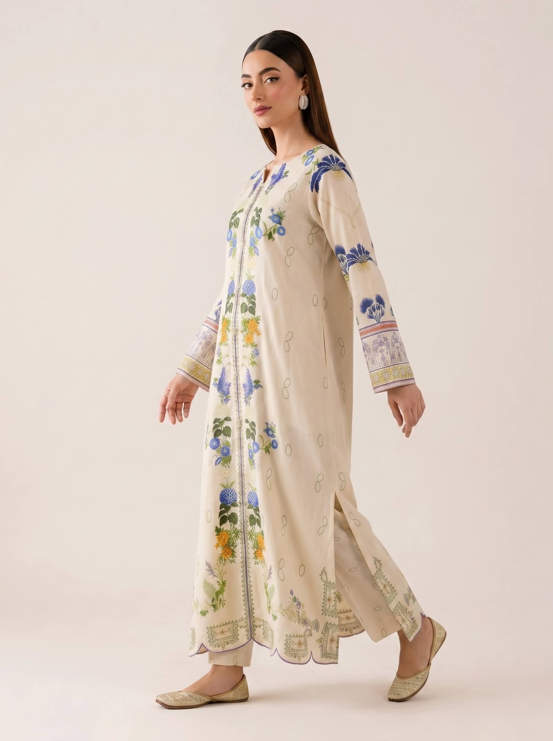A woman modeling a long cream-colored embroidered kurta with blue, green, and yellow floral patterns, paired with matching trousers and beige flat shoes, set against a solid light beige background.