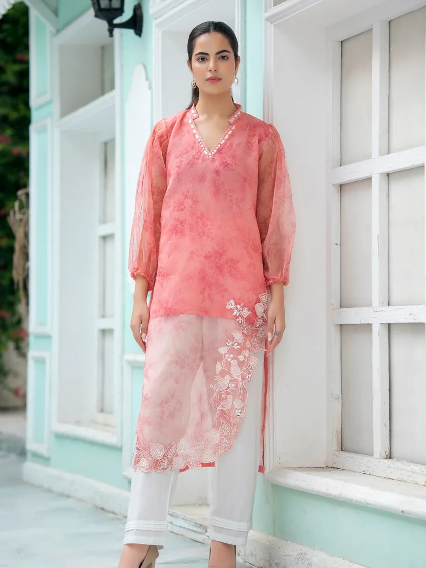 A woman models a three-piece traditional Indian outfit consisting of a coral pink printed kurta, a matching sheer embroidered dupatta, and white pants, standing in front of a white-framed window on a light mint green wall.