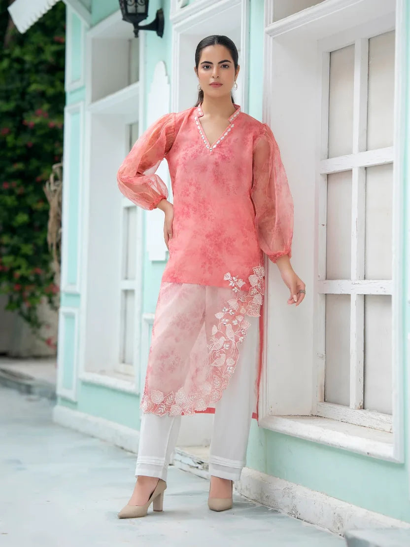 A woman models a three-piece peach pink and white printed ethnic suit consisting of a sheer long-sleeve kurta with floral embroidery, white pants, and beige heels, standing in front of a mint green building with white-framed windows.