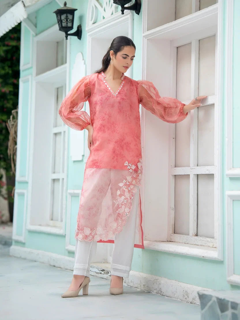 A woman models a peach-pink printed kurta with white pants and beige heels, standing in front of a mint green building with white-framed windows.