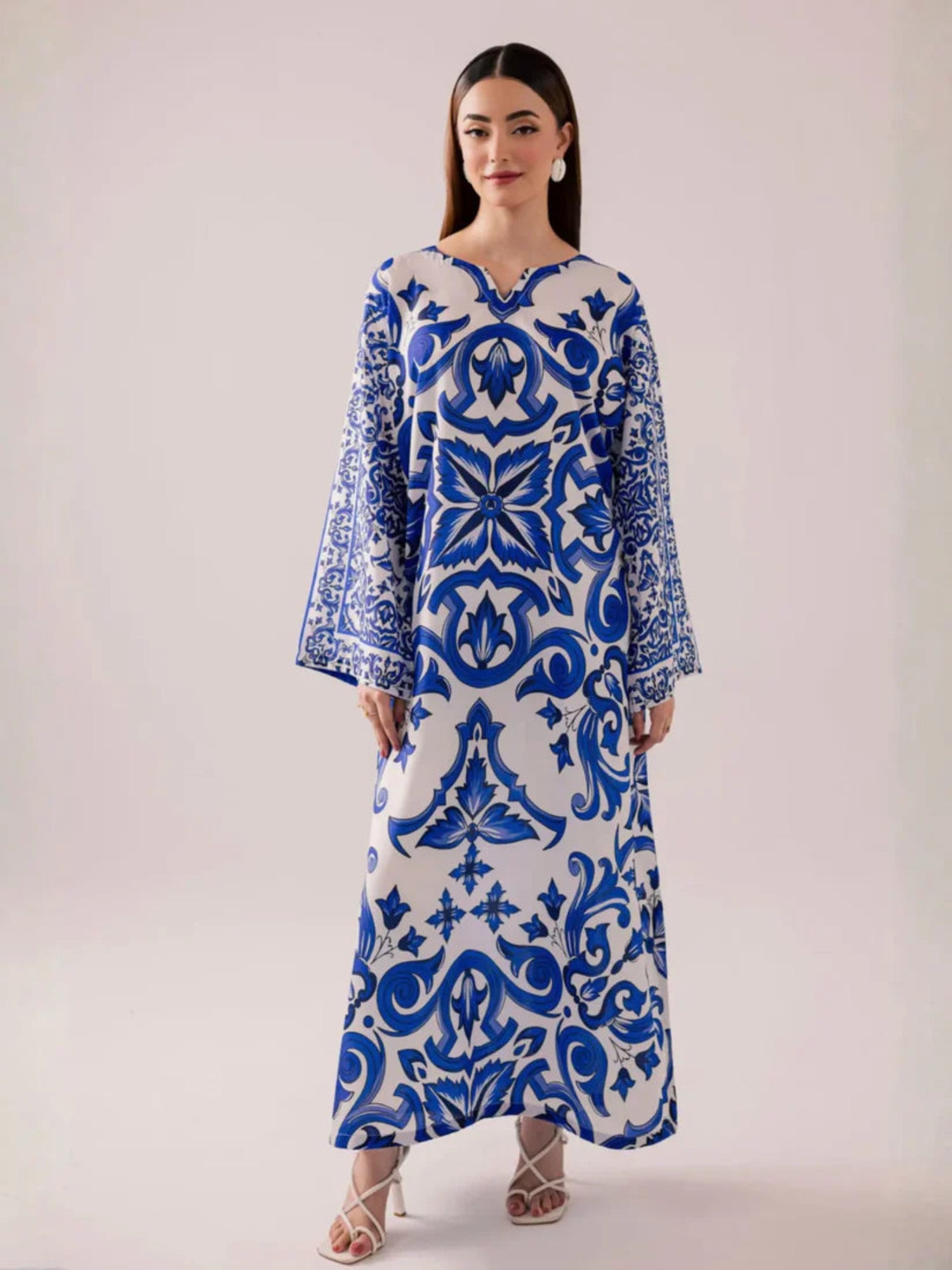 A woman modeling a long-sleeved maxi dress with a blue and white traditional damask pattern, standing against a solid light beige studio background.