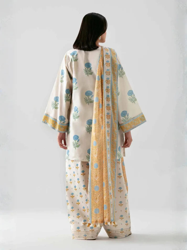 A model shown from the back wearing a traditional three-piece salwar kameez suit in cream with blue floral prints and a yellow-gold patterned dupatta, set against a plain light gray studio background.