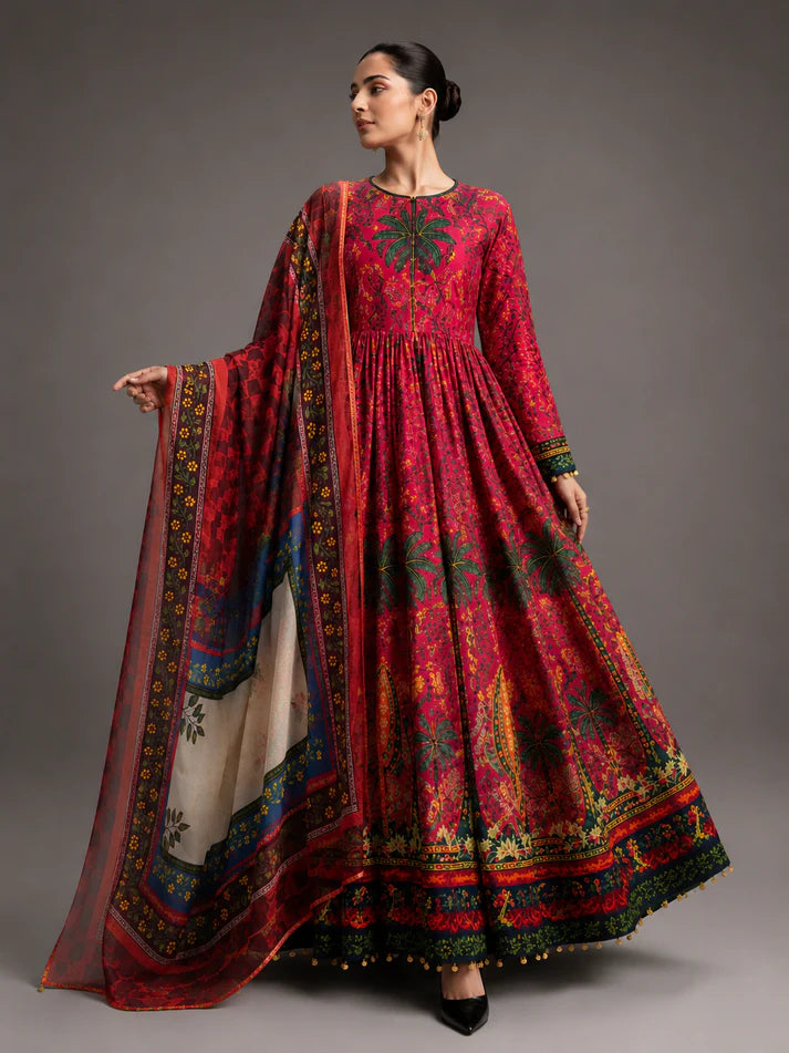 A model wearing a traditional pakistani Anarkali gown in red with intricate multicolor embroidery, paired with a matching sheer dupatta, set against a solid gray studio background.