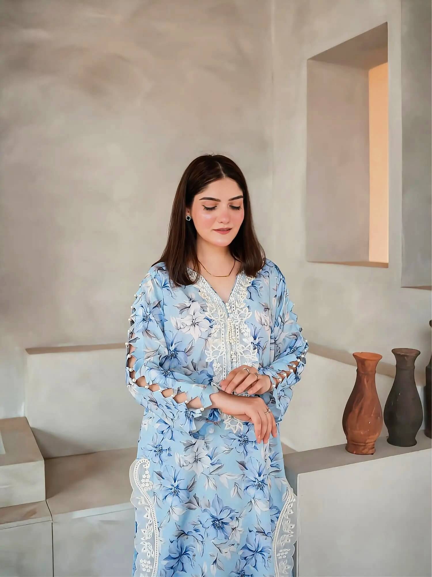 A woman wearing a light blue floral printed tunic with white lace trim stands in a minimalist interior setting with textured beige walls and decorative vases.