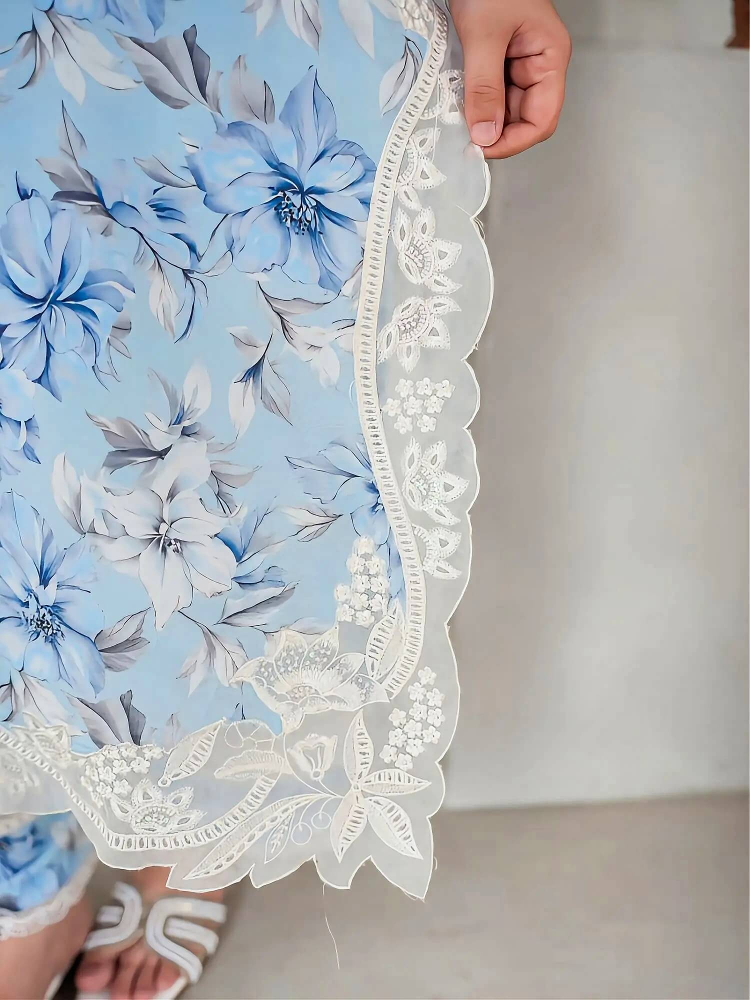 A close-up of a light blue floral fabric with white lace trim, held by a hand. The fabric features large blue and white flowers with gray leaves, and the lace has a scalloped edge with floral embroidery.
