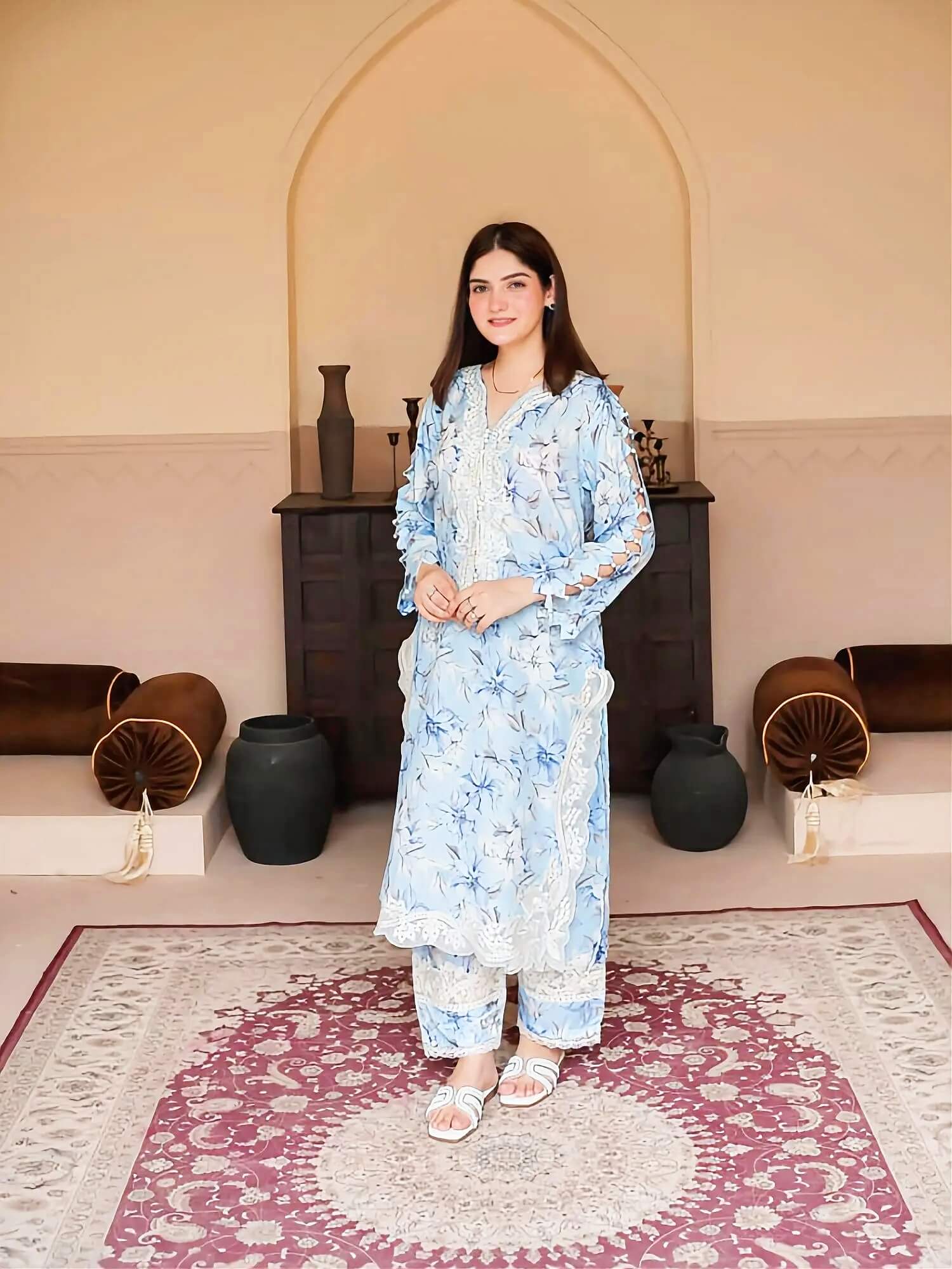 A woman stands indoors wearing a light blue and white floral printed kurta set with matching pants. The setting features a beige arched wall, a dark wooden cabinet, decorative vases, and a patterned rug.