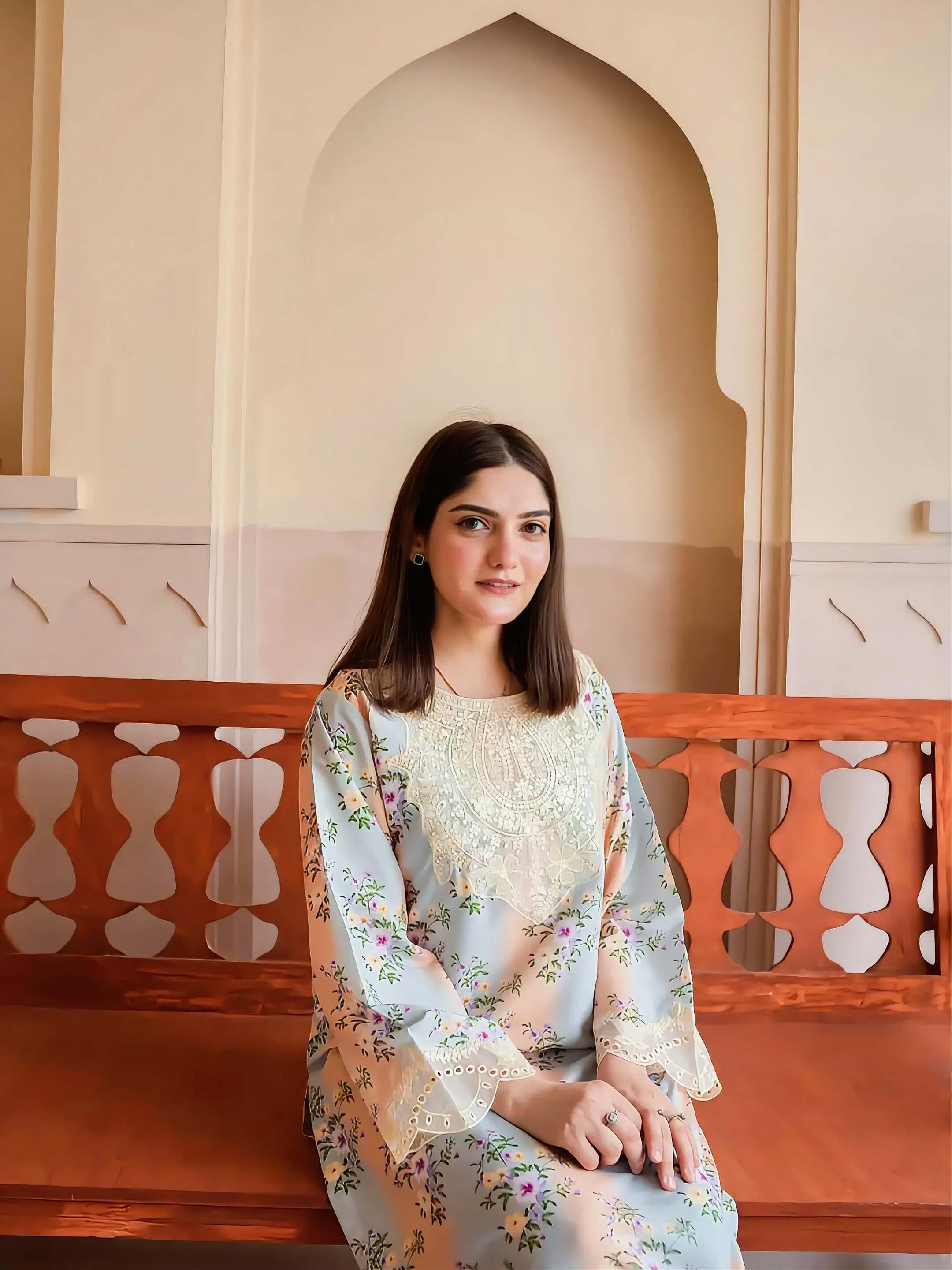 A woman with dark hair sits on a wooden bench in front of a cream-colored arched wall, wearing a light blue floral long-sleeve kurta with a cream embroidered yoke.