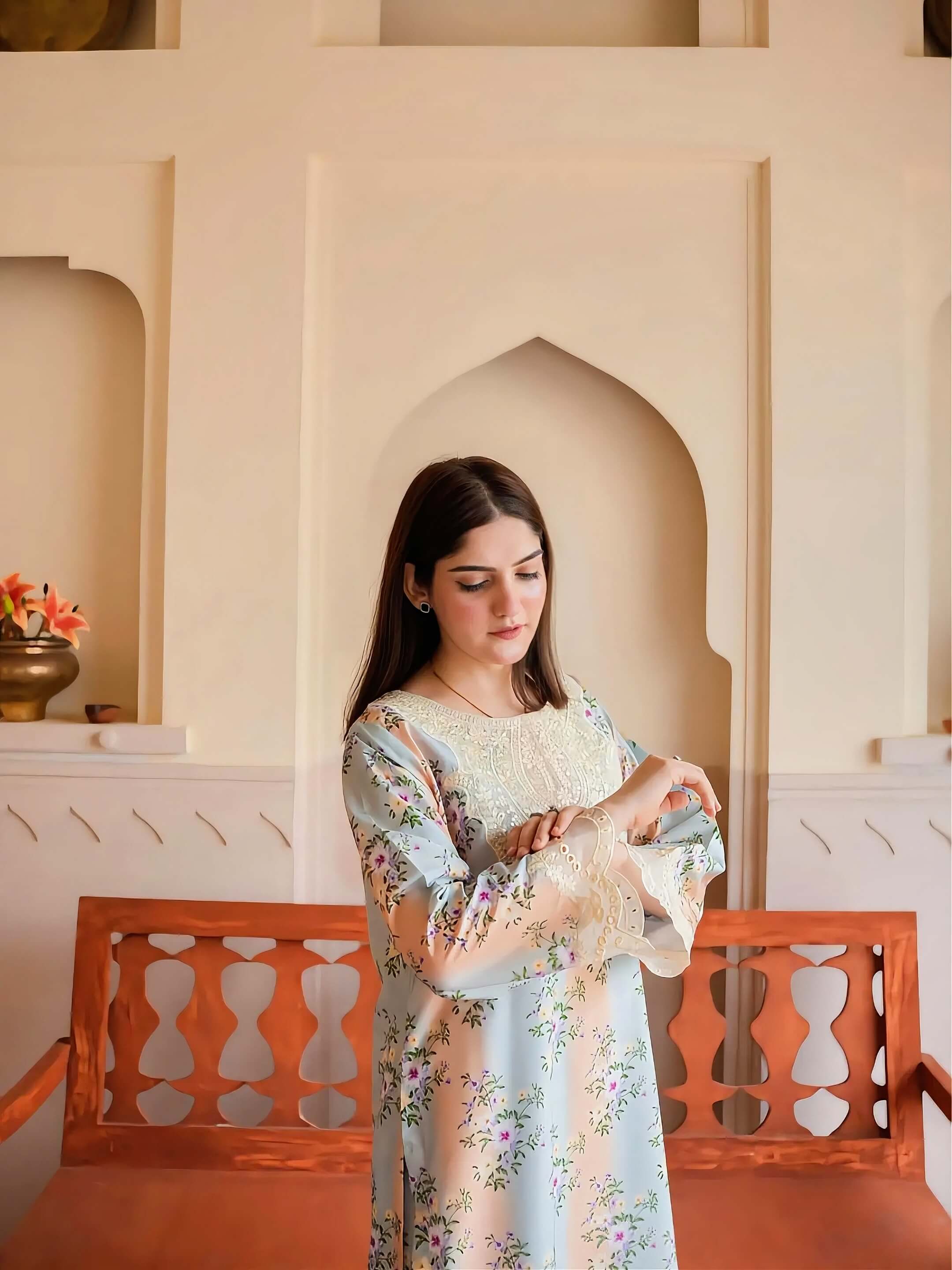 A woman wearing a light blue floral embroidered kurta stands in front of a cream-colored arched wall and a wooden bench.