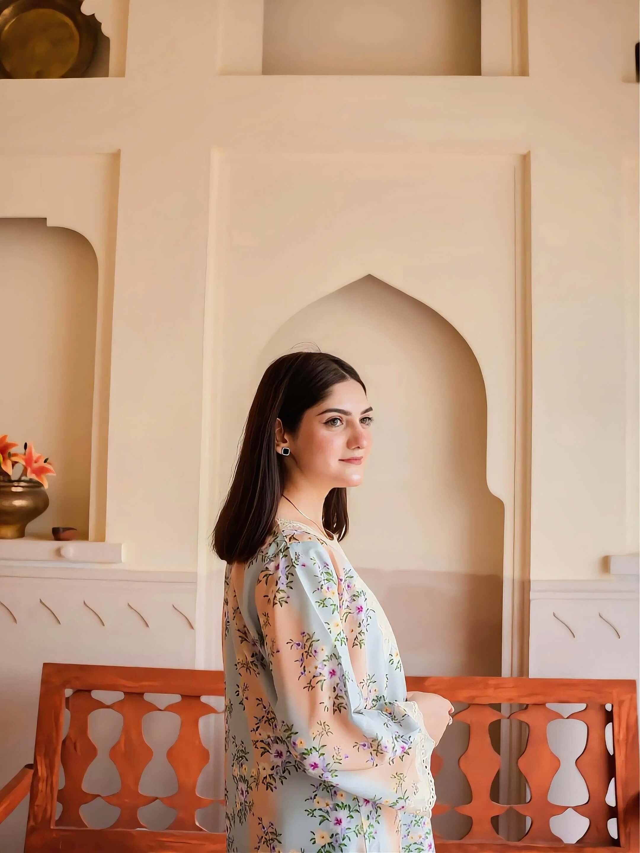 A woman with long dark hair stands in profile wearing a light blue floral tunic with beige sleeves, set against a cream-colored arched wall and a wooden bench.