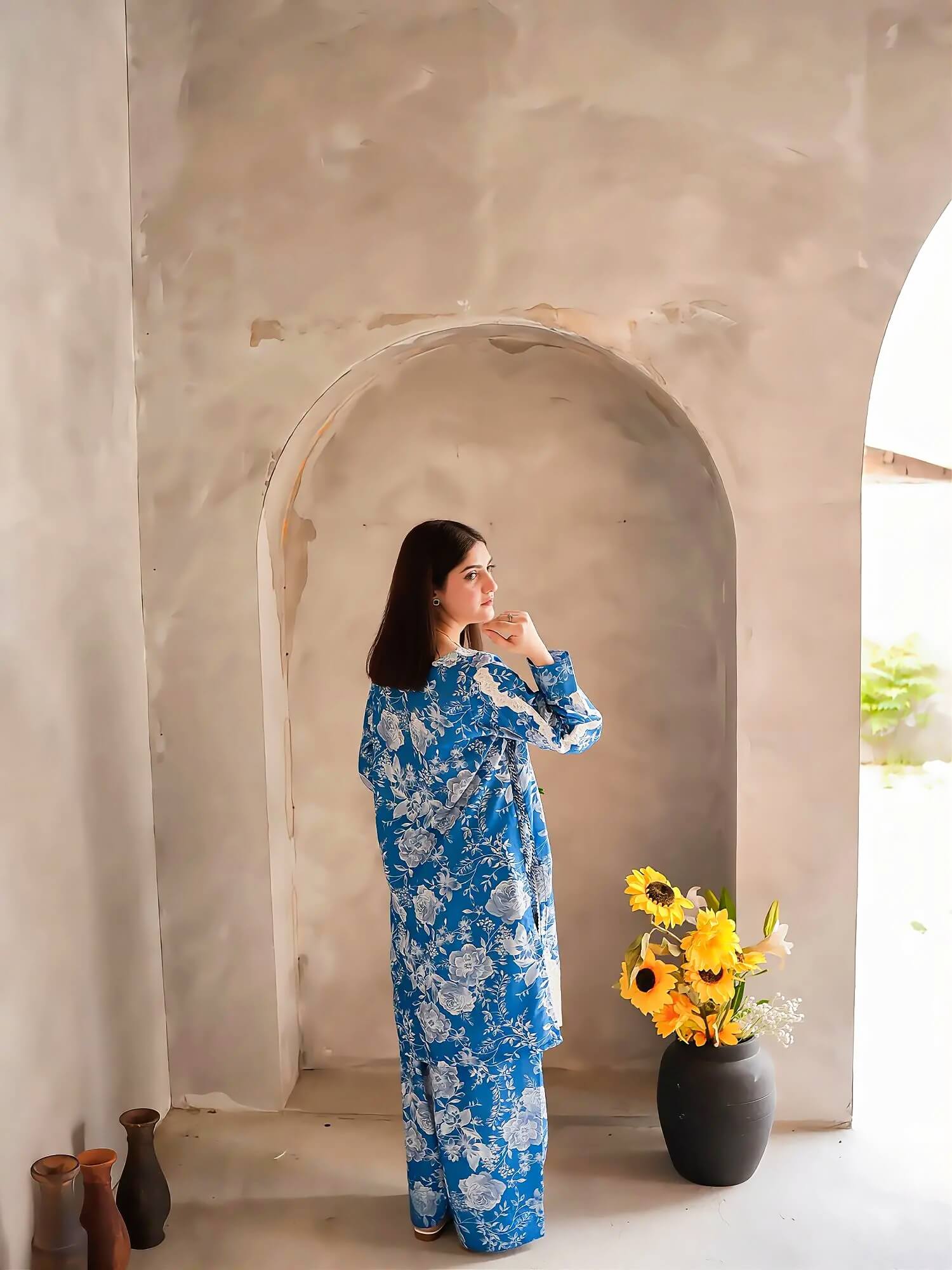 A woman wearing a blue and white floral long-sleeve jumpsuit stands in front of a plaster archway. A black vase with yellow sunflowers sits to her right, and a few small vases are on the floor to her left.