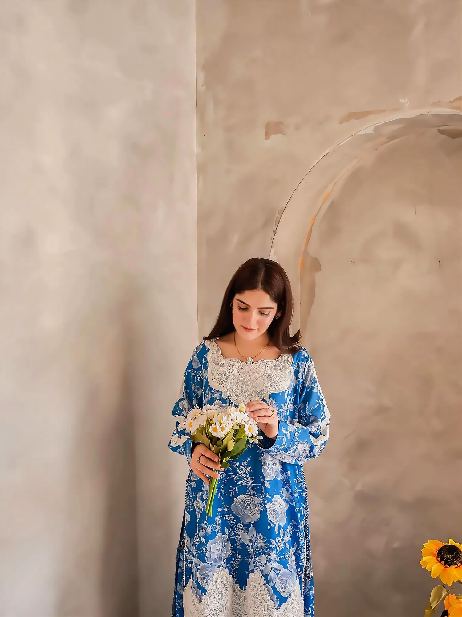 A woman wearing a blue and white floral print dress with lace trim, holding a bouquet of white flowers, standing against a textured beige plaster wall with a large archway behind her.