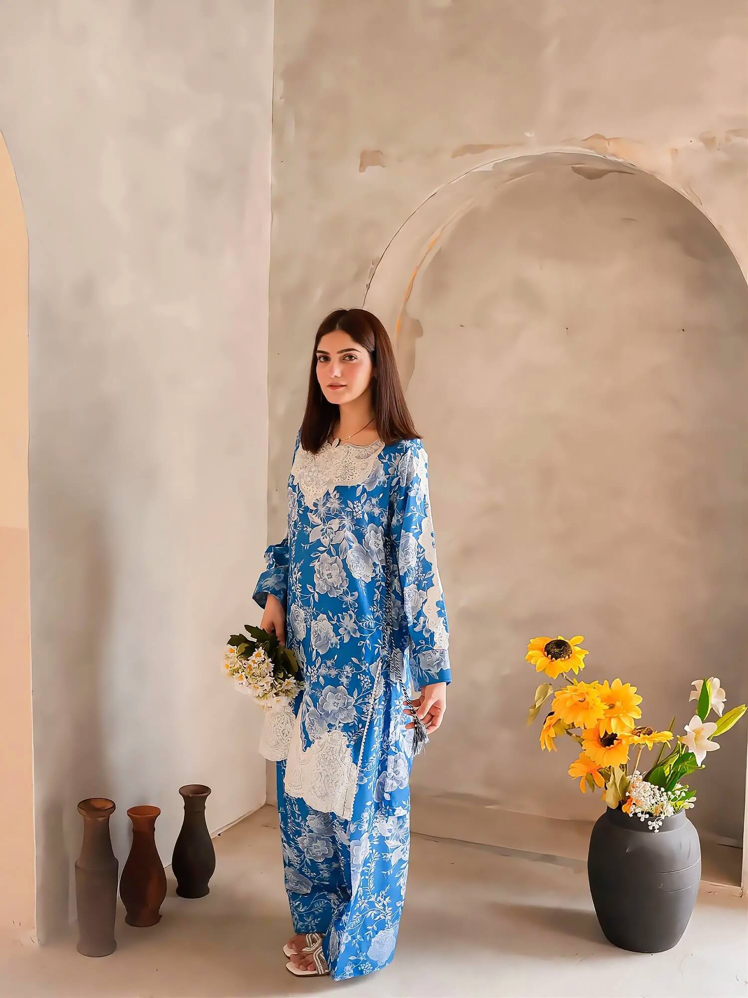 A woman stands in a rustic indoor setting wearing a blue and white floral print tunic and matching wide-leg pants. She holds a small bouquet of white flowers, with decorative vases and a large vase of sunflowers in the background.